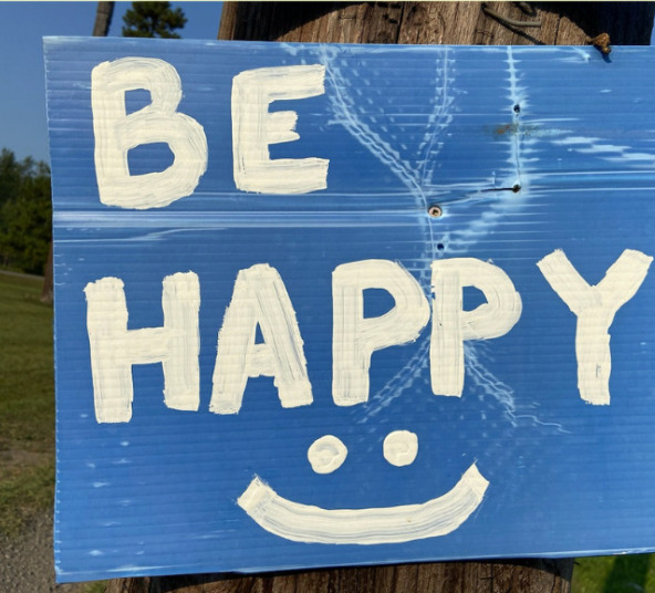A blue sign posted on an outdoor pole that says "Be Happy" in white letters with a smiley face